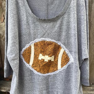 May include: A heather gray long-sleeve shirt with a scoop neckline and an Old Navy label. The center features an appliqué of a brown football with a textured pattern, white fabric laces, and a decorative white lace trim border around the edge.