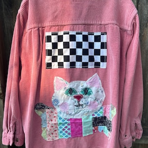 May include: Pink corduroy shirt with a patchwork cat design on the back. The cat has green eyes and a pink nose. A black and white checkered patch is above the cat. The collar has a black and white checkered pattern.