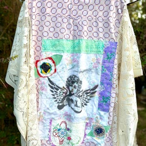 May include: A button-down shirt featuring a light pink and purple geometric pattern, cream-colored lace sleeves, and a central patchwork panel. The panel displays a printed cherub, floral fabric sections, a red flower applique, and embroidery.