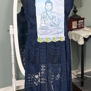 May include: Navy blue sleeveless dress with intricate lace detailing. The back features a rectangular patch with a light blue illustration of a seated Buddha, framed by blue and green floral accents. The dress is displayed on a black mannequin.