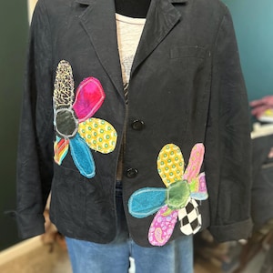 May include: Black blazer with two large, colorful flower appliqués. The flowers feature various patterns and colors, including leopard print, pink, yellow, and blue. The blazer has a classic lapel and button closure.