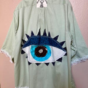 May include: A light green pinstriped button-down shirt featuring a large fabric eye appliqué on the back. The eye has a dark blue iris, a bright blue pupil, and white sclera. The shirt has white lace trim on the sleeves and hem and a label reading The House of Upcycle.