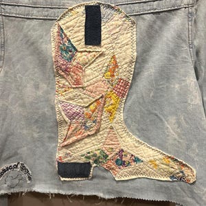 May include: A light blue denim jacket with a patchwork boot design on the back. The boot is made of various fabrics in shades of pink, yellow, and blue. The jacket has a frayed hem and a small embroidered patch that says "Lots of Trouble".