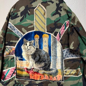 May include: Camouflage jacket with a large patch featuring a cat illustration. The jacket has various fabric patches in different colors and patterns, including stripes and floral designs. The cat is gray and white, sitting in front of a blue background with architectural elements.