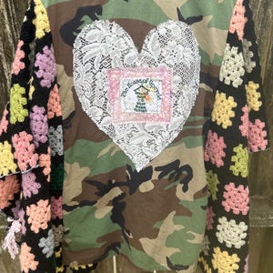 May include: Camouflage jacket with crochet sleeves and a large white lace heart on the back. The heart has a pink square with the text "The House of Upcycle" and a small illustration. The sleeves are black with colorful crochet squares.