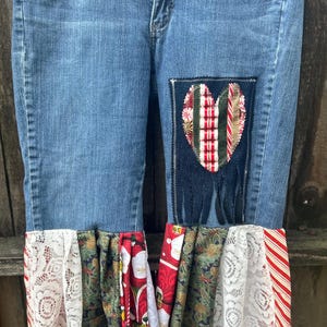 May include: Blue denim jeans with a heart-shaped patch and decorative fabric accents. The patch features a heart design with red and green stripes. The bottom of the jeans have a variety of fabrics, including white lace, Christmas-themed prints, and red and white stripes.