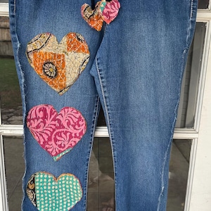 May include: Blue denim jeans with four patchwork hearts sewn on the front. The hearts are made of different fabrics with colorful patterns.