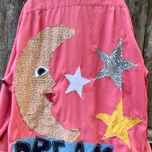 May include: A coral pink button-down shirt featuring large fabric appliqués on the back. The design includes a textured tan crescent moon with a face, a white star, a silver sequined star, a yellow star, and a blue fabric strip with the word DREAM.