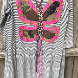 May include: A long-sleeved dress with a black and white striped pattern. The dress features a large butterfly design on the back, with pink, leopard print, and camouflage fabric accents. The butterfly has a decorative trim.