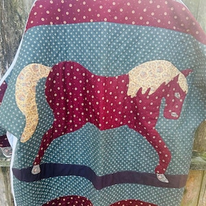 May include: Quilted jacket featuring a horse applique. The jacket showcases a patchwork design with burgundy, teal, and patterned fabrics. The horse is burgundy with a patterned mane and tail. The jacket has a relaxed fit and a unique, handcrafted aesthetic.