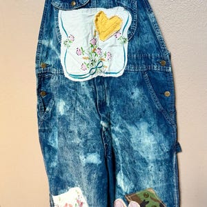 May include: Acid-wash blue denim overalls featuring a bib pocket with a white fabric patch displaying a yellow heart and floral embroidery. The legs are decorated with various fabric patches including floral patterns, a pink heart, and camouflage.