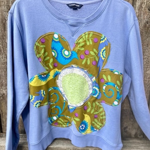 May include: A light blue Lands' End sweatshirt with a large, appliqued flower design. The flower features olive green, blue, and yellow patterned fabric with bird and leaf motifs. The center of the flower has a green patch with a white lace trim.