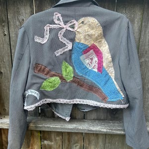 May include: Gray blazer with a decorative bird applique on the back. The bird is made of patterned fabric and lace, perched on a brown branch with green leaves. A pink lace bow and the word "upcycle" are also visible.