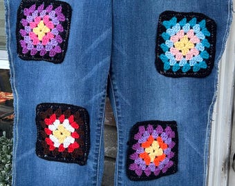 May include: Blue denim jeans with colorful crocheted squares sewn on the front. The squares are in various colors, including pink, purple, yellow, blue, red, and orange.