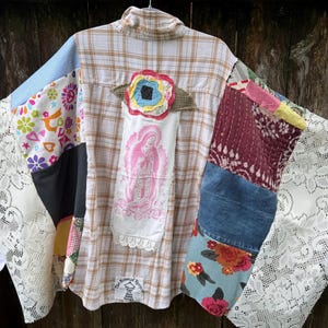 May include: A patchwork shirt featuring a tan and white plaid base with wide, white lace sleeves. The back displays a pink graphic of the Virgin of Guadalupe, a colorful fabric flower appliqué, and a small white label with text at the bottom hem.