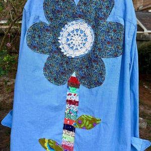May include: A light blue button-down shirt featuring a large appliquéd flower on the back. The flower petals are made of blue floral fabric with a white crocheted doily center. The stem is a vertical strip of multicolored fabric scraps.