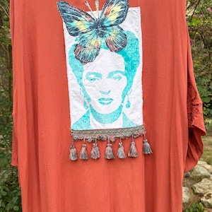 May include: A rust-colored kimono featuring a rectangular white patch on the back. The patch displays a teal-toned portrait of a person with a large, colorful butterfly positioned above the head. A decorative trim with small tassels hangs below.