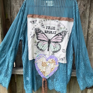May include: A teal-colored, long-sleeved top with a lace overlay and bell sleeves. The front features a white patch with a butterfly design and the words "Free Spirit." A patchwork heart is attached below.