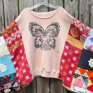 May include: A light pink sweatshirt featuring a large black butterfly graphic with the text Sunset Dream Music Festival. The garment is constructed with wide, patchwork-style sleeves made from various colorful fabrics, including red patterned textiles, blue floral squares, and white lace accents.