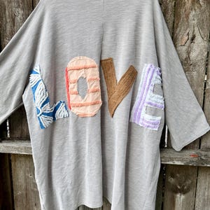 May include: A light gray ribbed cardigan featuring large, textured fabric letters on the back that spell out the word LOVE. Each letter is a different color and pattern, including blue and white, peach, brown, and light purple with frayed edges.