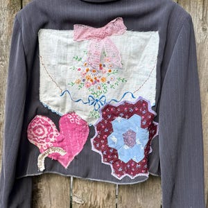 May include: A charcoal gray pinstriped blazer featuring a large white fabric patch on the back with floral embroidery and a pink lace bow. Below are a pink heart-shaped fabric appliqué and a multi-colored hexagonal patchwork design.