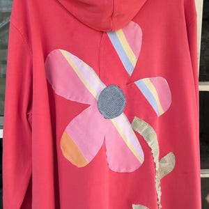 May include: A coral-colored hooded sweatshirt with a large flower applique on the back. The flower has pink, yellow, and blue striped petals and a gray center. The hood lining is striped.