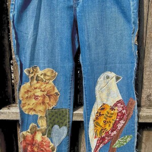 May include: Blue denim jeans with floral and bird appliqués. The jeans feature a decorative bird perched on a branch and floral patches. The bottom hems are trimmed with a brown tassel fringe. The jeans are labeled "Lee Classic Fit".