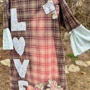 May include: Brown plaid upcycled shirt dress with floral and "LOVE" appliqués. The dress features bell sleeves with a light blue floral pattern and a faded red and brown plaid design. The text "The House of Upcycle" is visible on a patch.