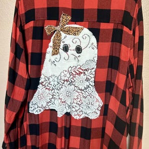 May include: Red and black plaid flannel shirt with a white ghost applique. The ghost has black eyes, a leopard print bow, and a lace bottom. The shirt has a button-down collar and long sleeves.