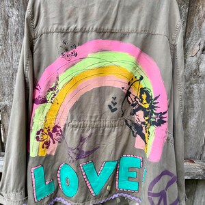 May include: An olive green utility jacket featuring a hand-painted design on the back. The artwork includes a vibrant rainbow, a black silhouette of a cherub with a bow and arrow, butterflies, birds, and the word LOVE in large teal letters.