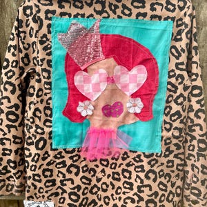 May include: A tan and black leopard print jacket with a whimsical patch featuring a cartoon face with pink hair, heart-shaped glasses, and a sequined crown. The patch is on a turquoise background.