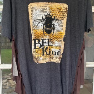 May include: Dark gray t-shirt with a large graphic of a bee and the words "BEE Kind" on a vintage paper background. The shirt has fringe detailing on the sides.