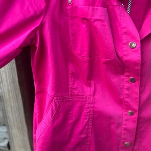 May include: A bright pink jacket with a collared neckline and button-down closure. The jacket features two front pockets and a single pocket on the chest. The sleeves are long, and the fabric appears to be a smooth, solid color.