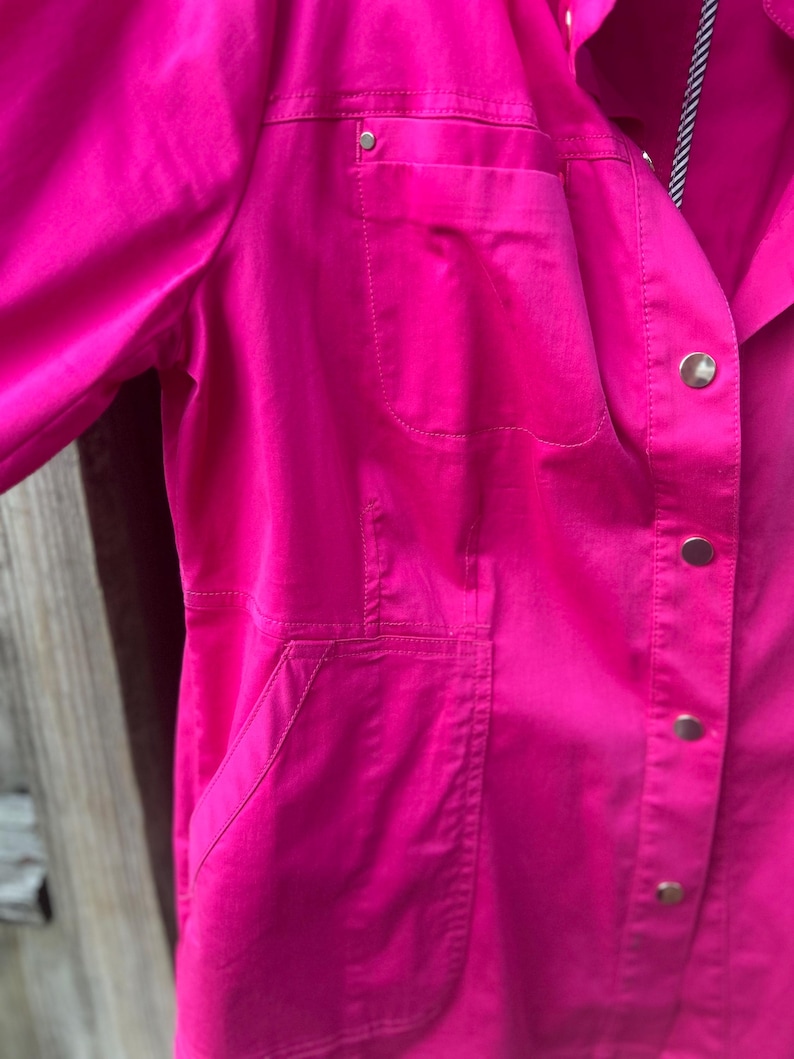 May include: A bright pink jacket with a collared neckline and button-down closure. The jacket features two front pockets and a single pocket on the chest. The sleeves are long, and the fabric appears to be a smooth, solid color.