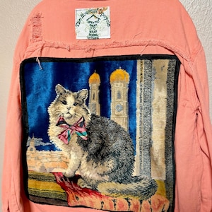 May include: A coral-colored denim jacket with a distressed hem and a patch featuring a cat wearing a bow tie. The jacket has a decorative patch with a cat design and the text "the house of upcycle".