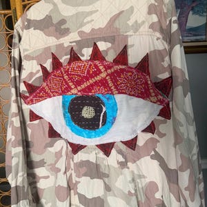 May include: A camouflage print jacket featuring a large, hand-stitched fabric eye appliqué on the back. The eye design includes a red patterned eyelid, a bright blue iris, and a dark pupil. A white rectangular label at the bottom reads The House of Upcycle.
