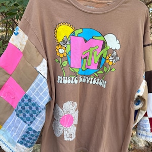May include: A brown long-sleeve t-shirt featuring a graphic of the MTV logo, a globe, a sun, and flowers with the text MUSIC TELEVISION. The sleeves are constructed from a patchwork of various fabrics, including pink, blue, and white patterns.