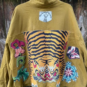 May include: A mustard yellow jacket featuring a large central tiger patch, a floral patch on the left, a butterfly patch on the right, and a smaller patterned patch. A white label at the top reads The House of Upcycle Life is too short to wear boring clothes.