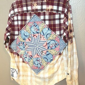 May include: A red and white plaid flannel shirt with a patchwork quilt design on the back. The quilt is made of various shades of blue, pink, and yellow fabric. The shirt has been bleached and has a distressed look.