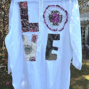 May include: A white long-sleeved shirt featuring the word LOVE appliqued on the back. The letter L is dark brown with a pattern, the O is a round floral lace doily, the V is cream with colorful floral motifs, and the E is a green camouflage fabric.