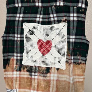 May include: A sleeveless plaid shirt with a crocheted square patch featuring a red heart in the center. The shirt has a bleached effect and a brown and white plaid pattern on the bottom half.