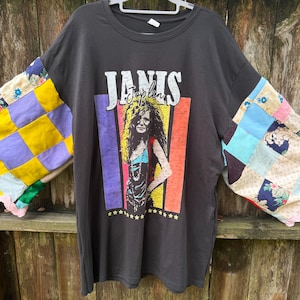 May include: A charcoal gray t-shirt featuring a graphic of Janis Joplin with the text "JANIS" and "Joplin" above her image. The shirt has wide, bell-shaped sleeves made from a colorful patchwork of floral and solid fabric squares with pink scalloped trim.