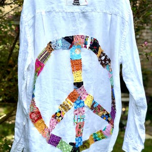 May include: A white long-sleeve button-down shirt featuring a large peace sign on the back constructed from various colorful patterned fabric patches. A label at the neck reads The House of Upcycle, Life is too short to wear boring clothes.