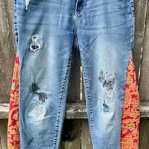 May include: Blue denim jeans with distressed details and colorful fabric panels on the sides. The jeans feature painted butterfly and floral designs, adding a unique, artistic touch to the garment.