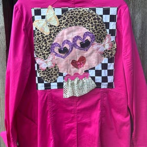 May include: A bright pink jacket with a whimsical design on the back. The design features a face with heart-shaped glasses, a leopard print background, and butterfly accents. The jacket has a classic button-up style.