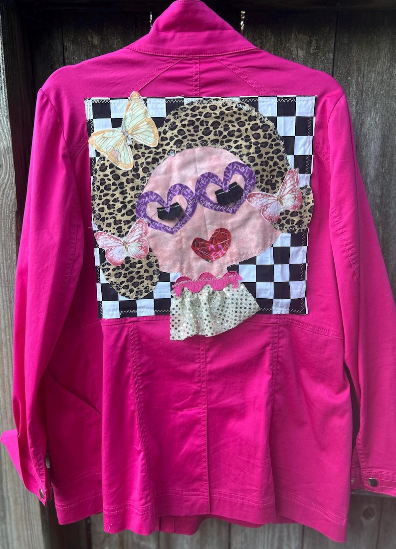 May include: A bright pink jacket with a whimsical design on the back. The design features a face with heart-shaped glasses, a leopard print background, and butterfly accents. The jacket has a classic button-up style.
