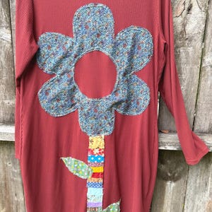 May include: A long-sleeved, ribbed, brick-red dress featuring a large, six-petaled flower appliqué made of blue patterned fabric. The stem is constructed from a vertical strip of multicolored, patchwork fabric scraps with two small green leaves.