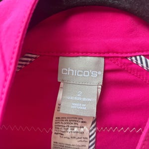 May include: Close-up of a bright pink Chico's shirt tag. The tag is white with black text, including the brand name, size, and fabric composition. The shirt is a vibrant fuchsia color, with a striped lining visible.