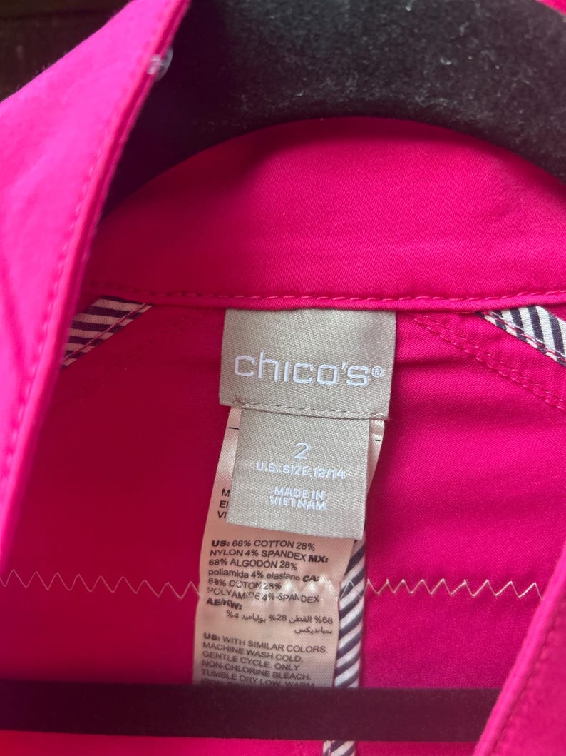 May include: Close-up of a bright pink Chico's shirt tag. The tag is white with black text, including the brand name, size, and fabric composition. The shirt is a vibrant fuchsia color, with a striped lining visible.