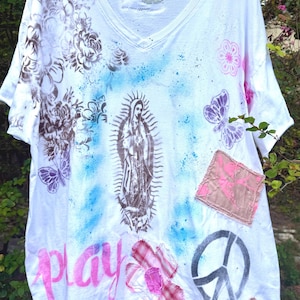 May include: A white V-neck t-shirt featuring a central brown graphic of the Virgin of Guadalupe, surrounded by blue spray paint, brown floral stencils, purple butterflies, a pink peace sign, a pink fabric patch, and the word play in pink.